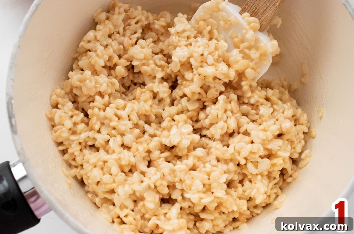A white pan filled with a warm, gooey Rice Krispie Treat mixture, ready for coloring and shaping, with a white wooden spoon resting in it.