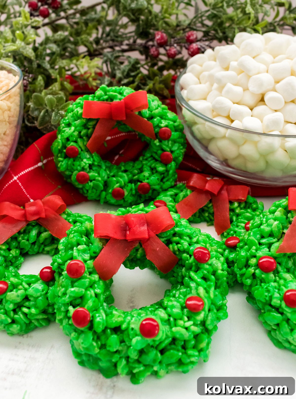 Six beautifully decorated Christmas Wreath Rice Krispie Treats resting on a white table, with glass bowls of Mini Marshmallows and Rice Krispies Cereal in the background.