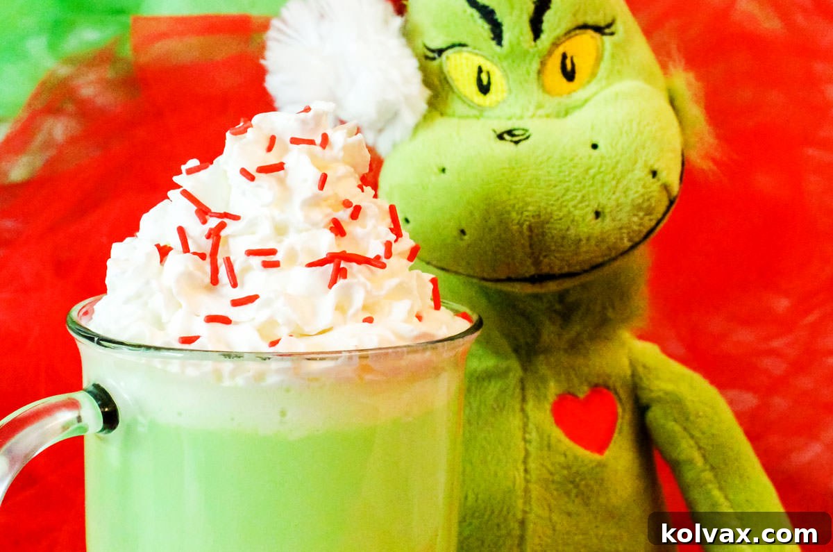 A glass mug overflowing with vibrant green Grinch Hot Vanilla Milk, generously topped with fluffy white whipped cream and festive red sprinkles, sits beside a cuddly Grinch plush toy. The scene evokes a cozy, Christmassy atmosphere perfect for holiday movie nights.