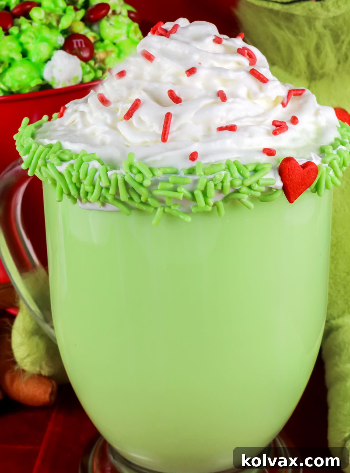 A delightful close-up of a mug filled with Grinch Hot Vanilla Milk, artfully topped with a swirl of whipped cream and a sprinkle of vibrant red sugar crystals, ready to be enjoyed as a festive Christmas drink.