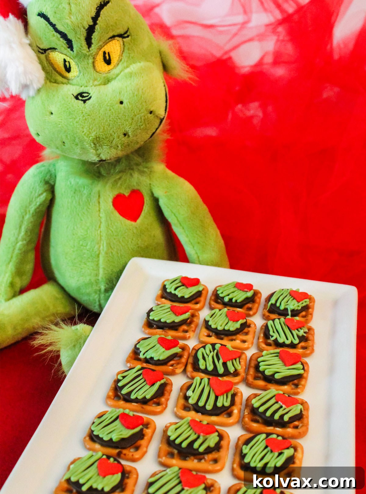 A batch of Grinch Pretzel Bites on a white serving platter sitting on a red table next to a Grinch stuffed animal, ready to be enjoyed.