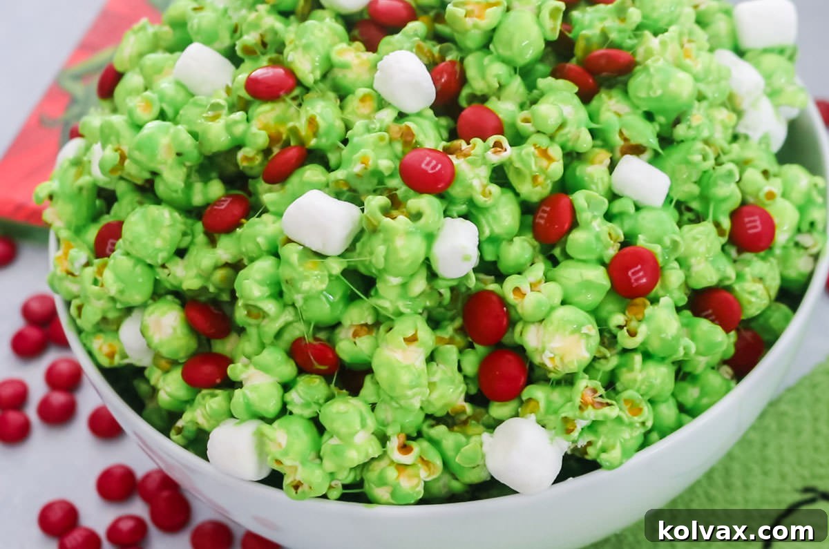 Closeup on a white serving bowl filled with Grinch Popcorn sitting on a grey table surrounded by Red M&M's and a grinch kitchen towel.