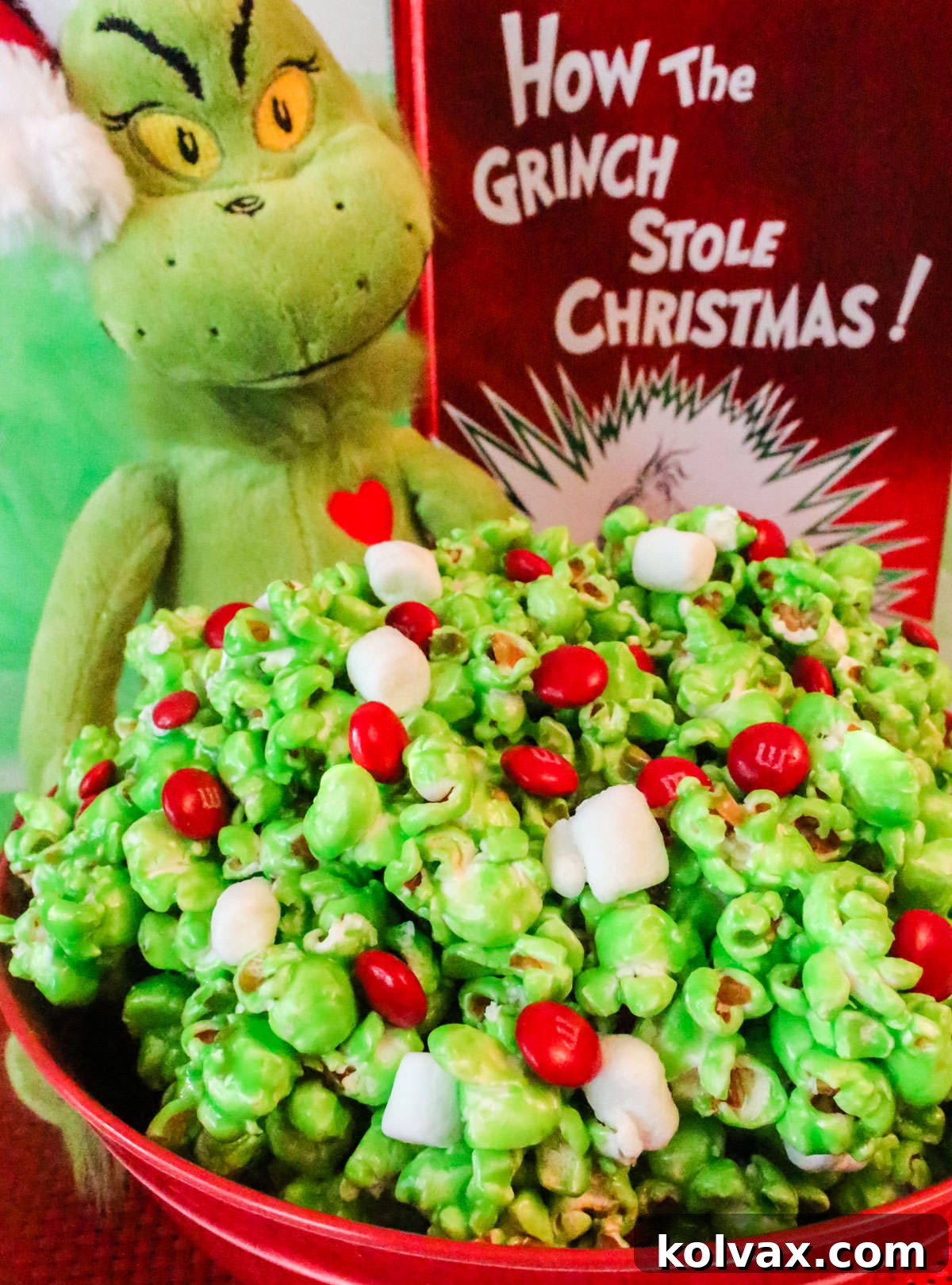 Closeup on a red bowl filled with Grinch Popcorn sitting in front of a Grinch stuffed animal and a copy of the book, How the Grinch Stole Christmas.
