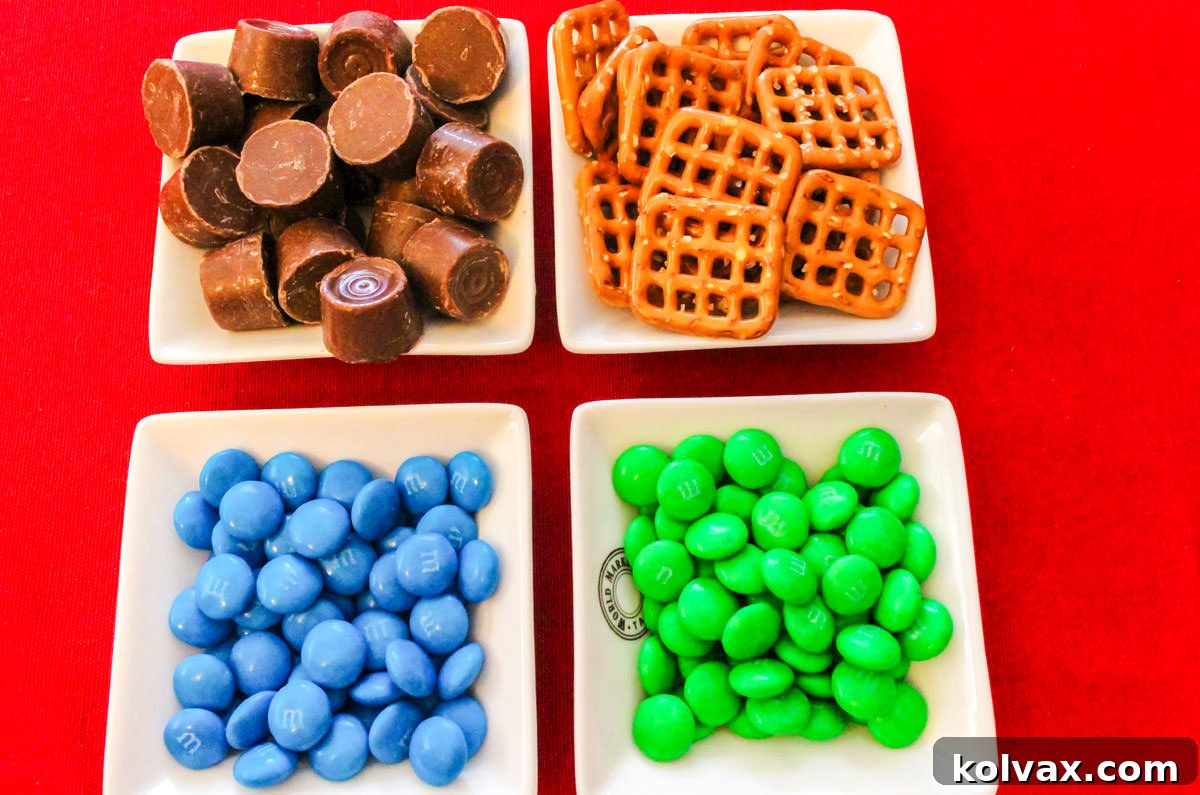 Four white ramekins neatly arranged on a red table, filled with a colorful assortment of blue and green M&M's, classic Rolo candies, and square Pretzel Snaps, showcasing all the ingredients needed for the recipe.