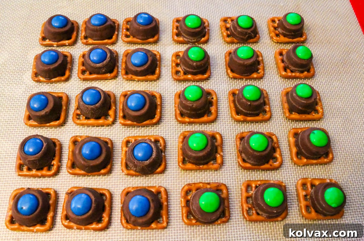 A vibrant batch of freshly made Seattle Seahawks Pretzel Bites, featuring blue and green M&M's on melted Rolo candies atop square pretzels, neatly lined up on a cookie sheet, ready for a game day party.