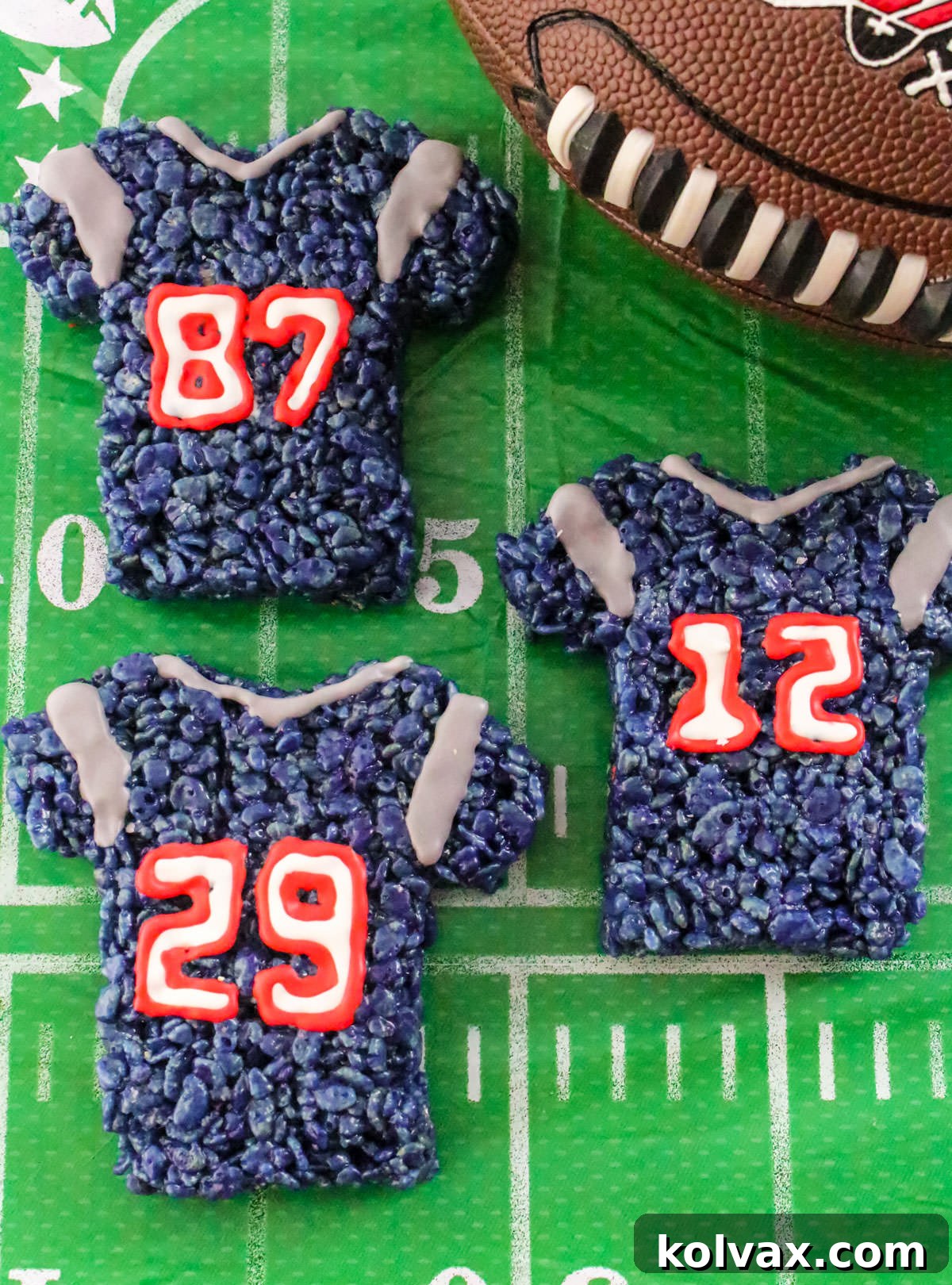 Touchdown Treats for Patriots Fans 5 An overhead view of three New England Patriots Rice Krispie Treats shaped like football jerseys, artfully arranged on a football-themed tablecloth beside a small football, showcasing festive game day desserts.