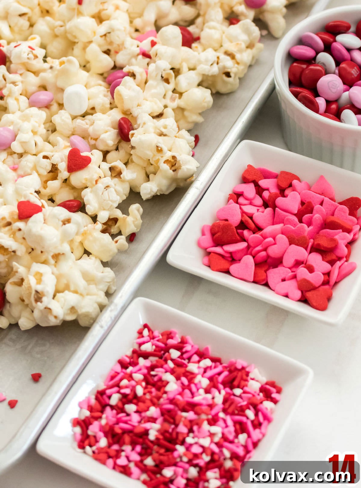 A close-up of three small white bowls, each filled with different Valentine's Day mix-ins: red and pink sprinkles, heart-shaped sprinkles, and colorful Valentine's M&M's. They are placed next to a cookie sheet covered with freshly made Valentine's Day Popcorn, ready for decoration.