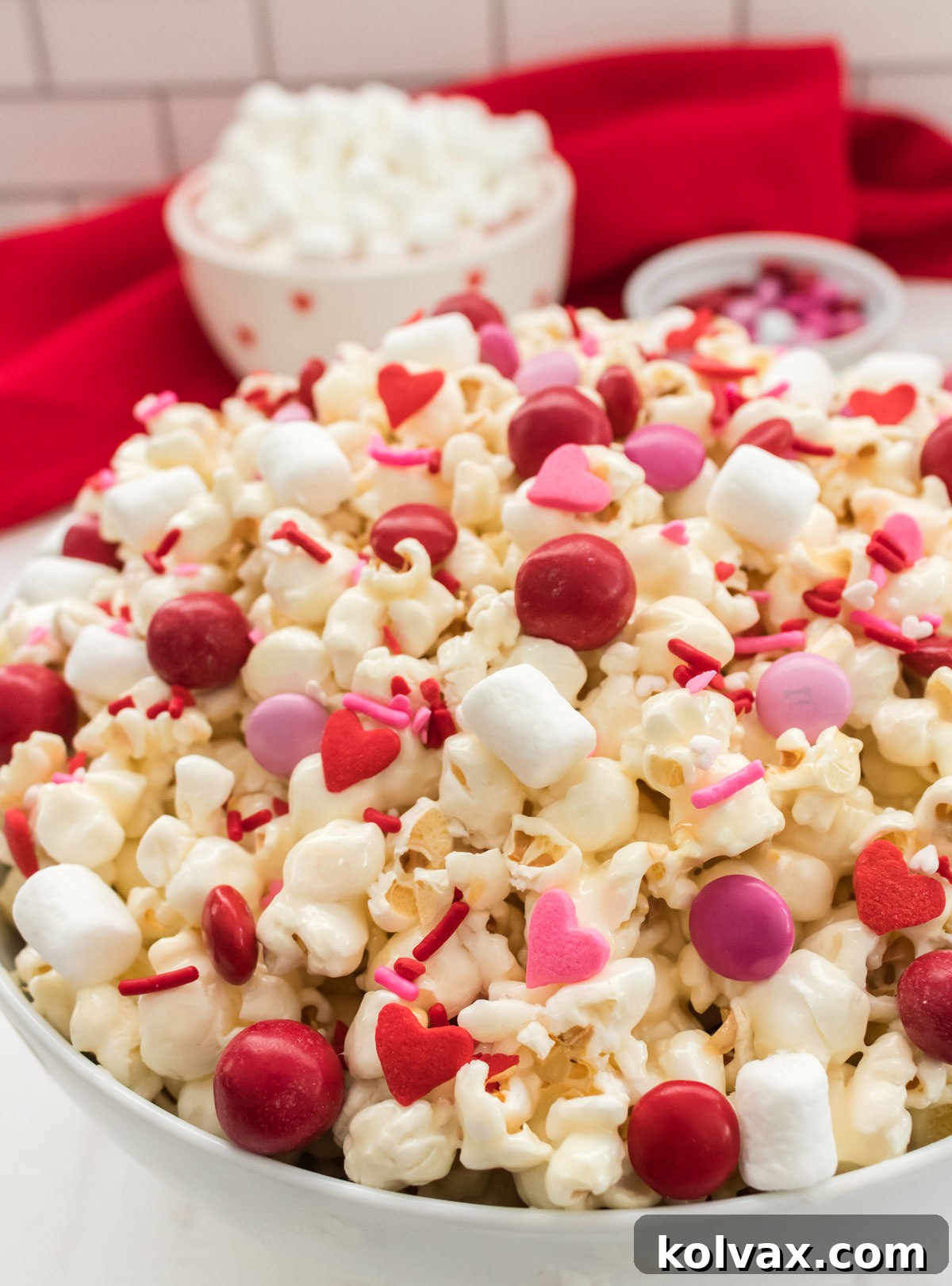 A beautifully arranged white bowl filled with Valentine's Day Popcorn, sitting on a pristine white table. In the background, slightly out of focus, are small bowls of mini marshmallows and colorful M&M's, emphasizing the ingredients used to create this festive treat.