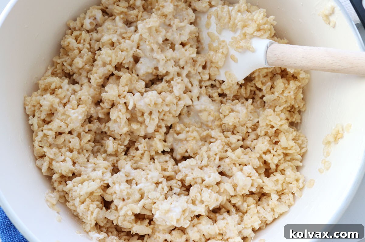 A white saucepan filled with a gooey, warm Rice Krispie Treat mixture, ready for cereal, being stirred with a wooden spatula.