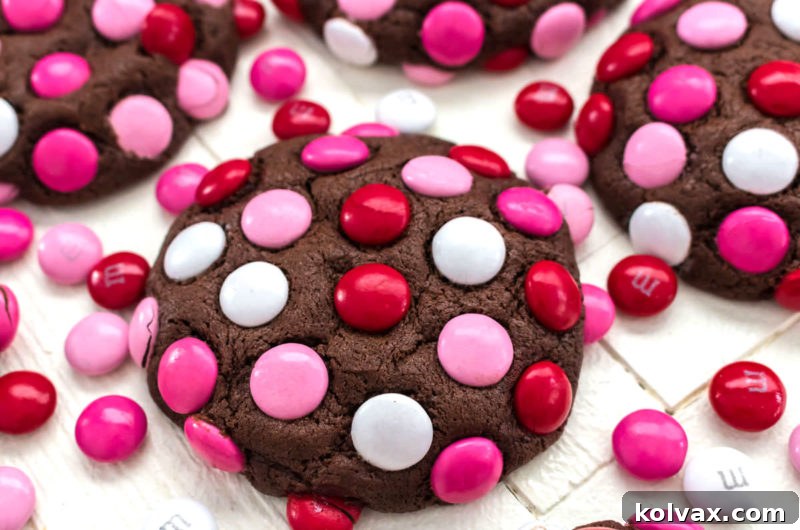 Colorful Valentine M&M Cookies 2 Close-up of a festive Valentine's Day M&M Cookie on a white surface, surrounded by loose red, bright pink, light pink, and white M&M's, highlighting its brownie-like texture.