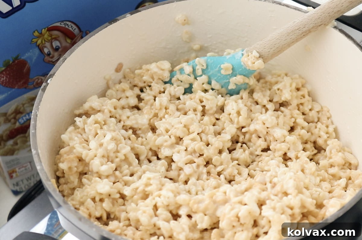A white saucepan filled with the warm, gooey marshmallow and butter mixture, ready for the Rice Krispie cereal, with a wooden spatula resting inside.