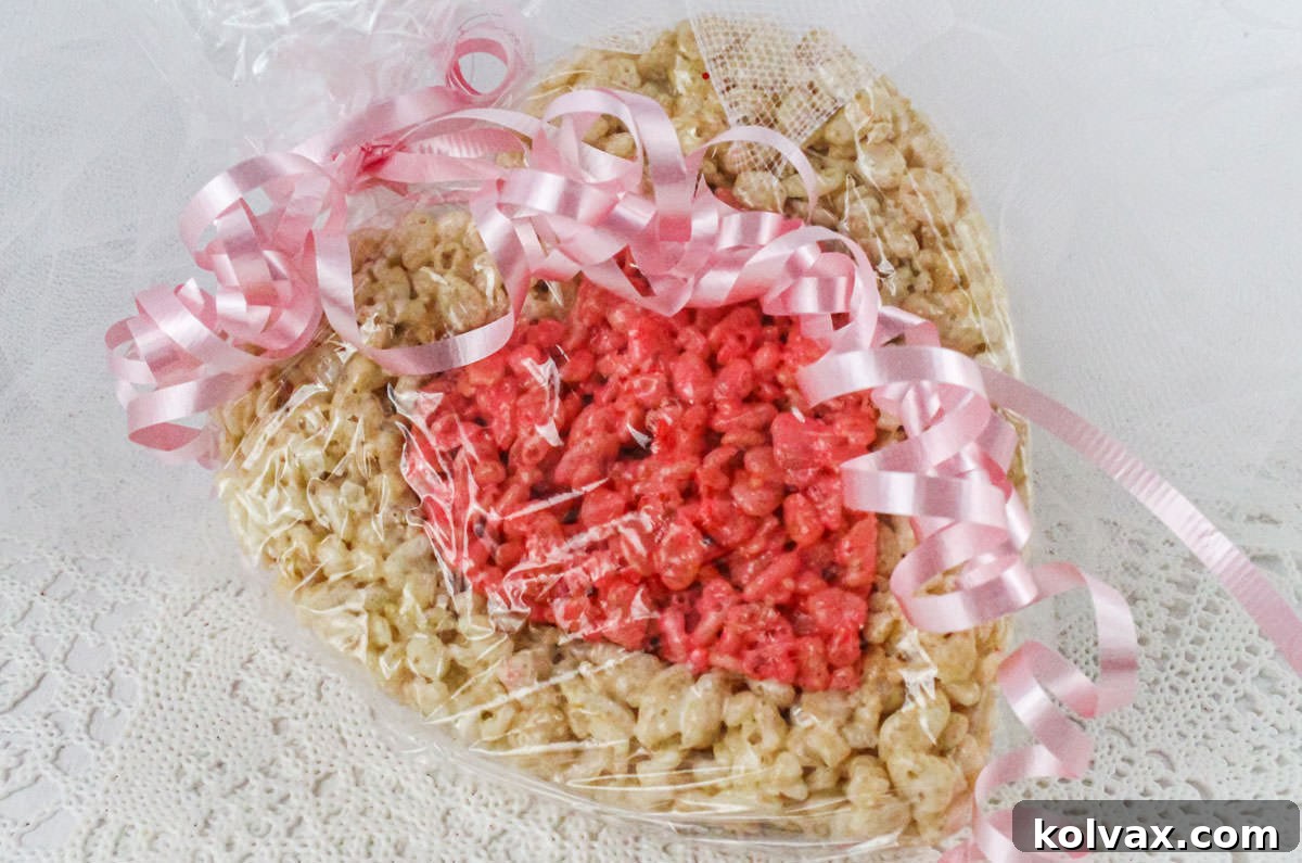 A charming pink and white Rice Krispie Treat heart elegantly wrapped in clear plastic wrap and tied with a delicate pink ribbon, ready to be presented as a thoughtful Valentine's gift.