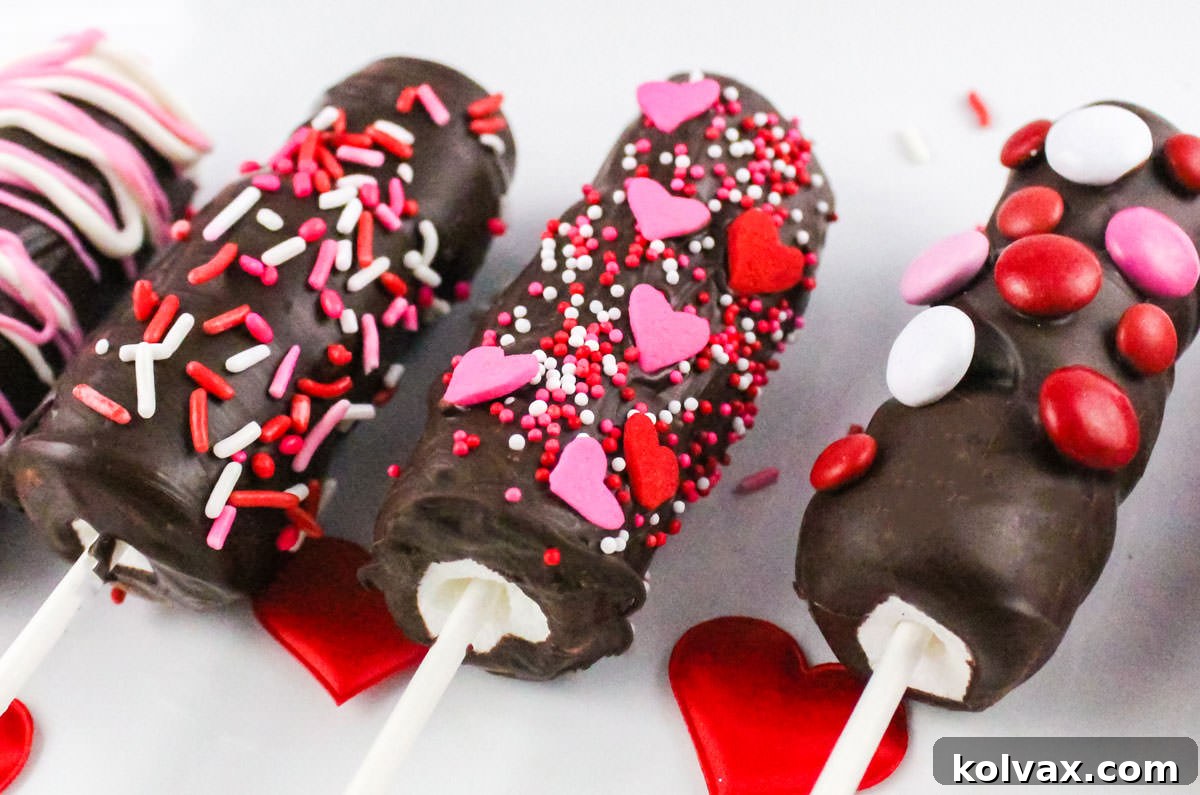 Closeup of three Valentine's Day Marshmallow Pops decorated with red, white and pink sprinkles, hearts and M&M's. These easy chocolate-covered marshmallow wands are perfect for Valentine's Day gifts or parties.