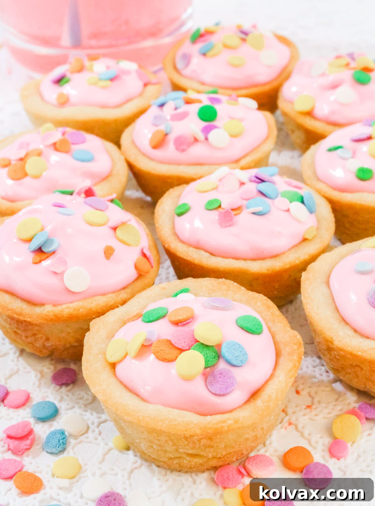Closeup on a batch of Pink Lemonade Cookie Cups sitting on a white table in front of a glass of Pink Lemonade, showcasing their readiness to serve and vibrant color.