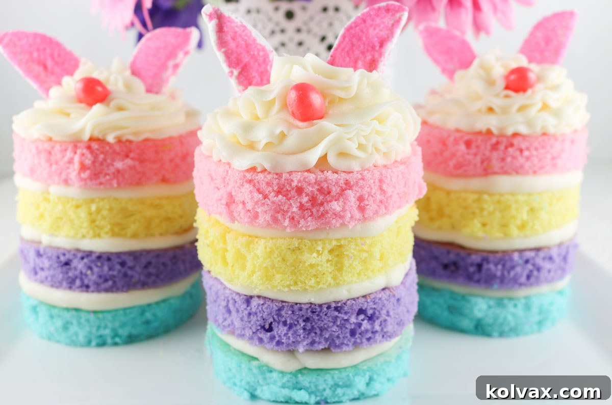 A close-up of three delightful Easter Bunny Mini Cakes, each with four pastel layers, creamy white frosting, pink marshmallow ears, and a pink jelly bean nose. They are artfully arranged on a white surface, with a white vase holding pink and purple daisies softly blurred in the background, creating a charming and festive scene.
