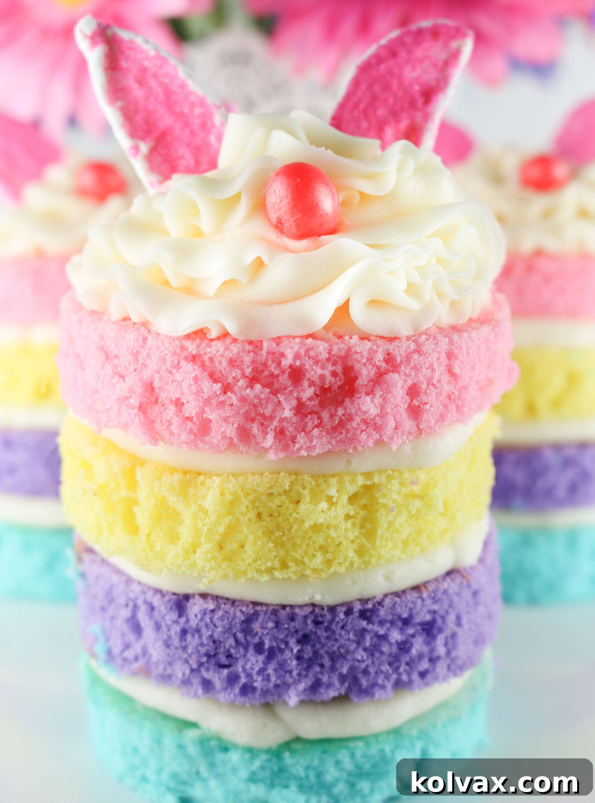 A close-up of a single Easter Bunny Mini Cake, perfectly assembled with its four pastel layers, white buttercream swirl, pink marshmallow ears, and a jelly bean nose. It sits on a white surface with two other bunny cakes softly blurred in the background, creating depth and focus on the featured dessert.