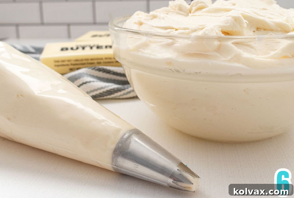A close-up of a clear glass bowl brimming with fluffy white homemade buttercream frosting, positioned next to a piping bag also filled with the creamy frosting, indicating readiness for cake decoration.