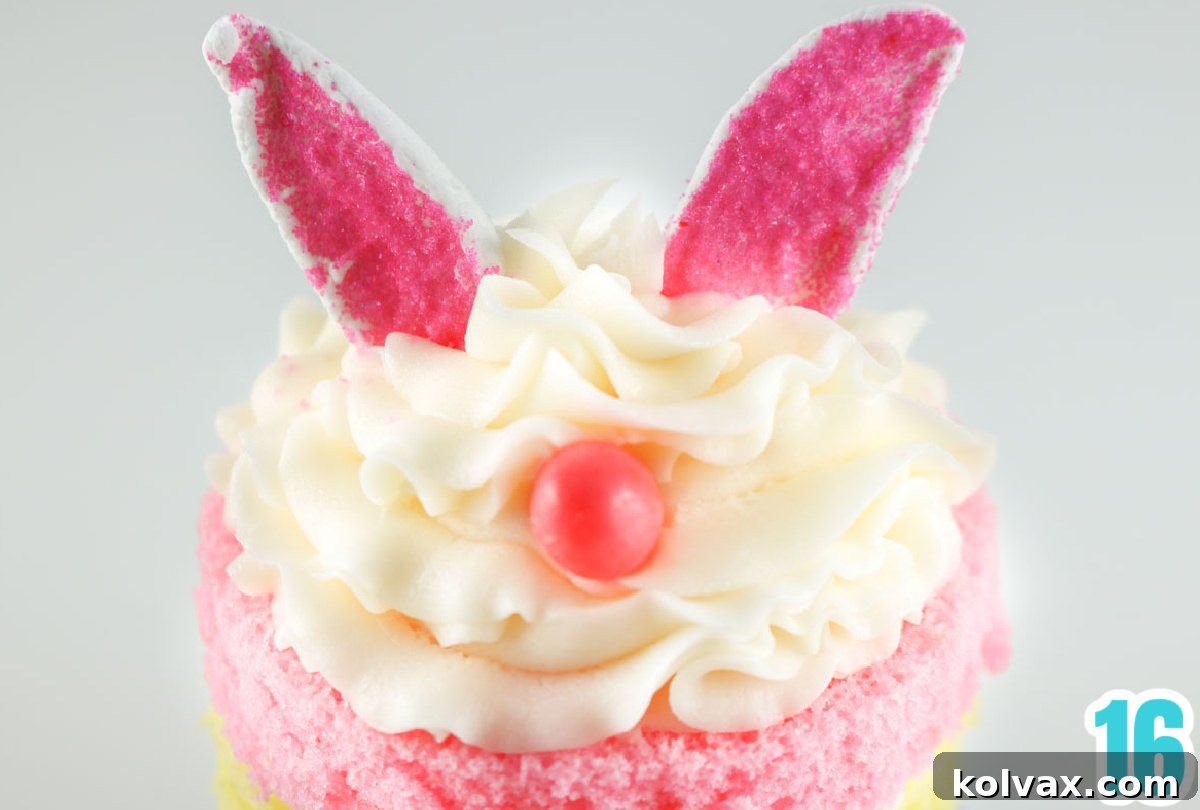 A close-up view of the top of an Easter Bunny Mini Cake, showcasing the fluffy white buttercream swirl, pink marshmallow bunny ears firmly set, and a small pink jelly bean serving as the cute bunny nose.