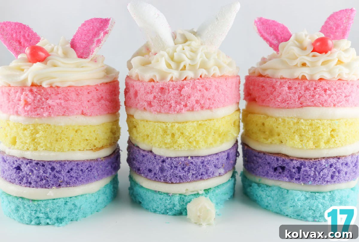 A close-up of three Easter Bunny Mini Cakes, turned to reveal their delightful frosting bunny tails on the back. The cakes are artfully arranged on a white surface, with soft light highlighting their pastel layers.