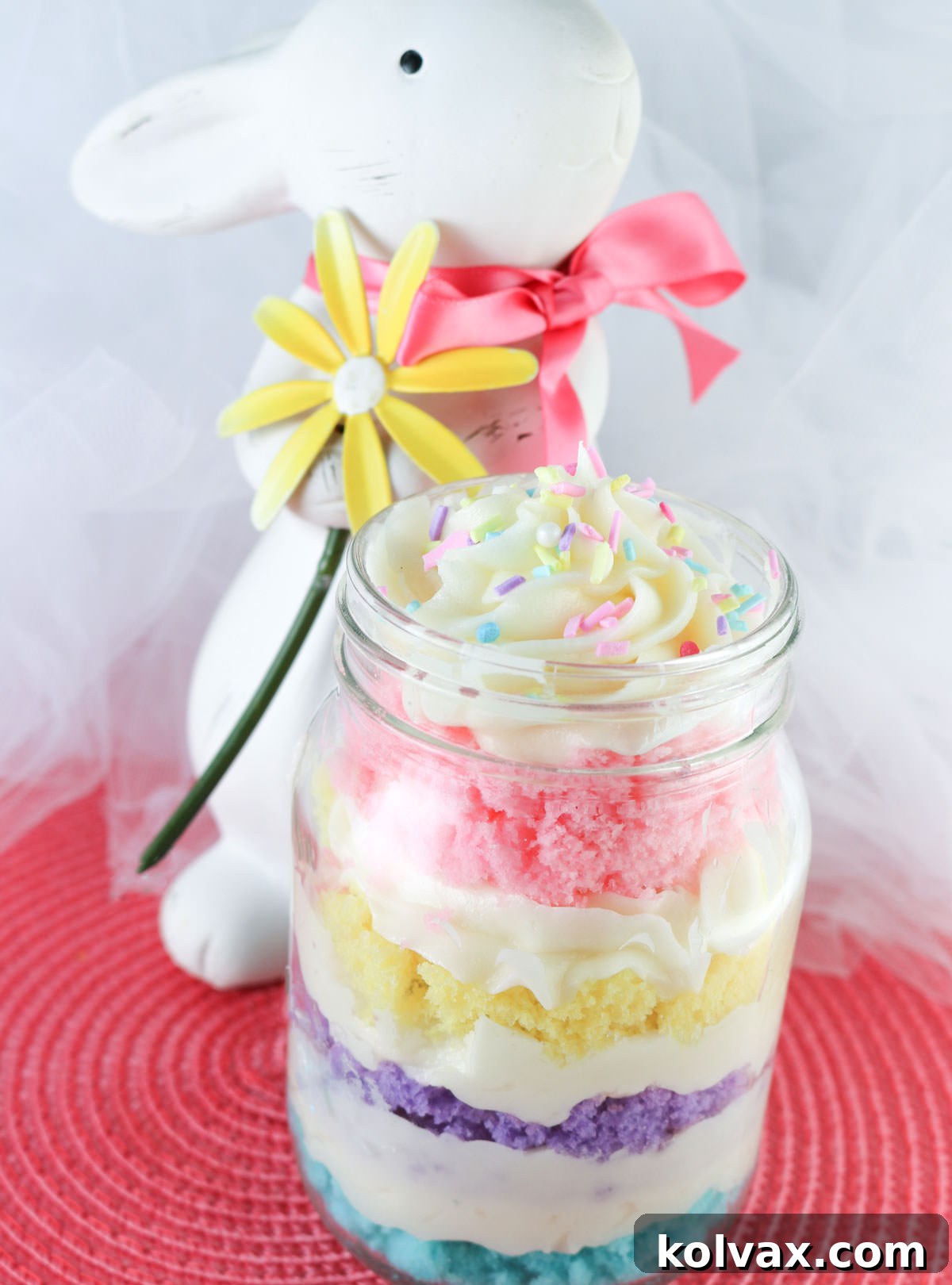 A charming close-up of a single Springtime Cupcake in a Jar, perfectly layered and topped, placed on a pink placemat beside a white Easter bunny decoration holding a yellow flower, setting a festive scene.