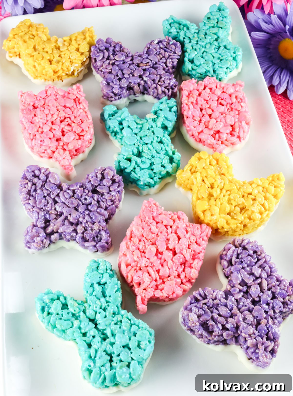 A large white platter covered with Blue, Purple, Pink and Yellow Rice Krispie Treats dipped in White Chocolate surrounded by pink and purple decorative daisies.