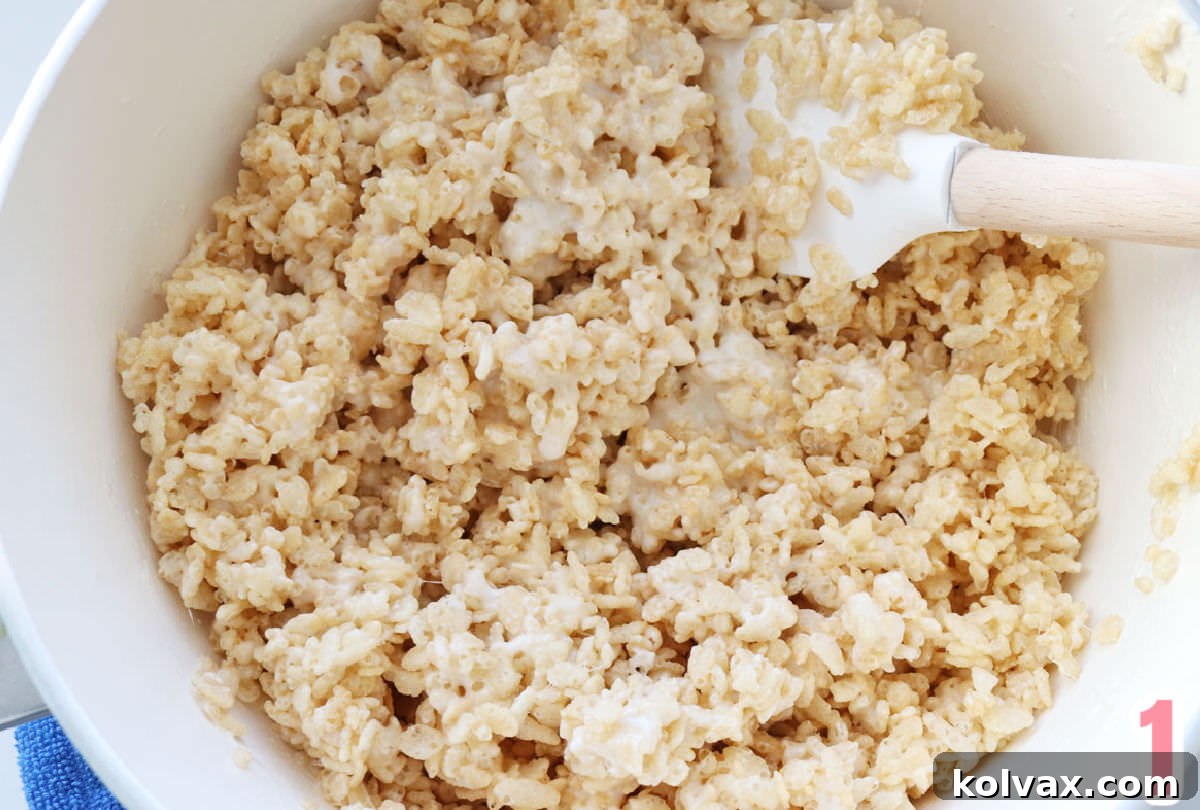A pristine white saucepan filled with a freshly made, warm Rice Krispie Treat mixture, ready for molding, accompanied by a wooden spatula.