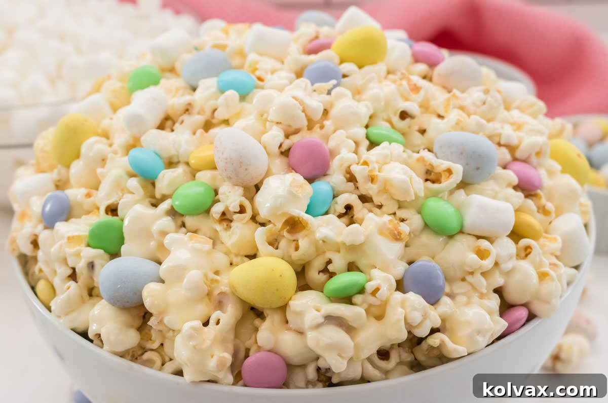 Close up on a white serving bowl filled with Easter Candy Popcorn sitting on a white table in front of a bowl of mini marshmallows and a pink table linen, highlighting the colorful candies and fluffy popcorn.