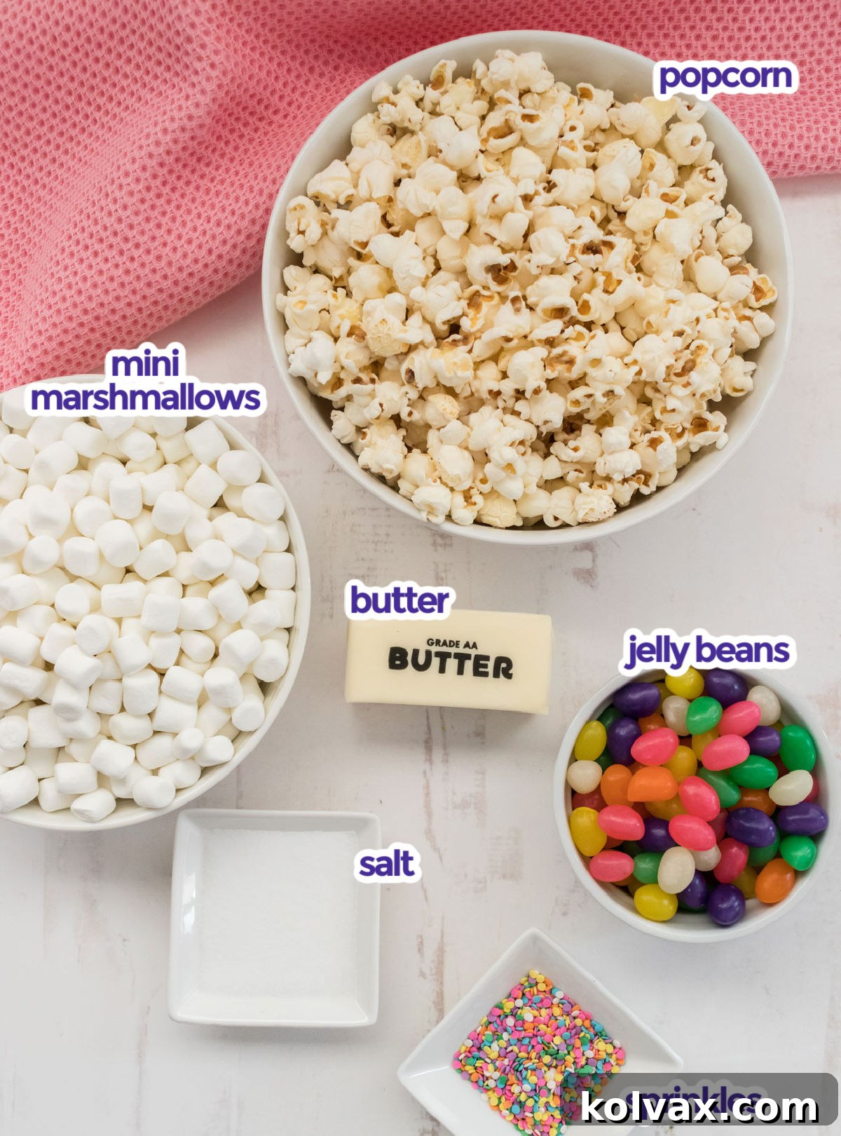 A beautifully arranged flat lay of all the fresh ingredients required to make Jelly Bean Popcorn, including fluffy popcorn kernels, miniature marshmallows, a stick of butter, a salt shaker, a generous assortment of colorful jelly beans, and festive Easter sprinkles.