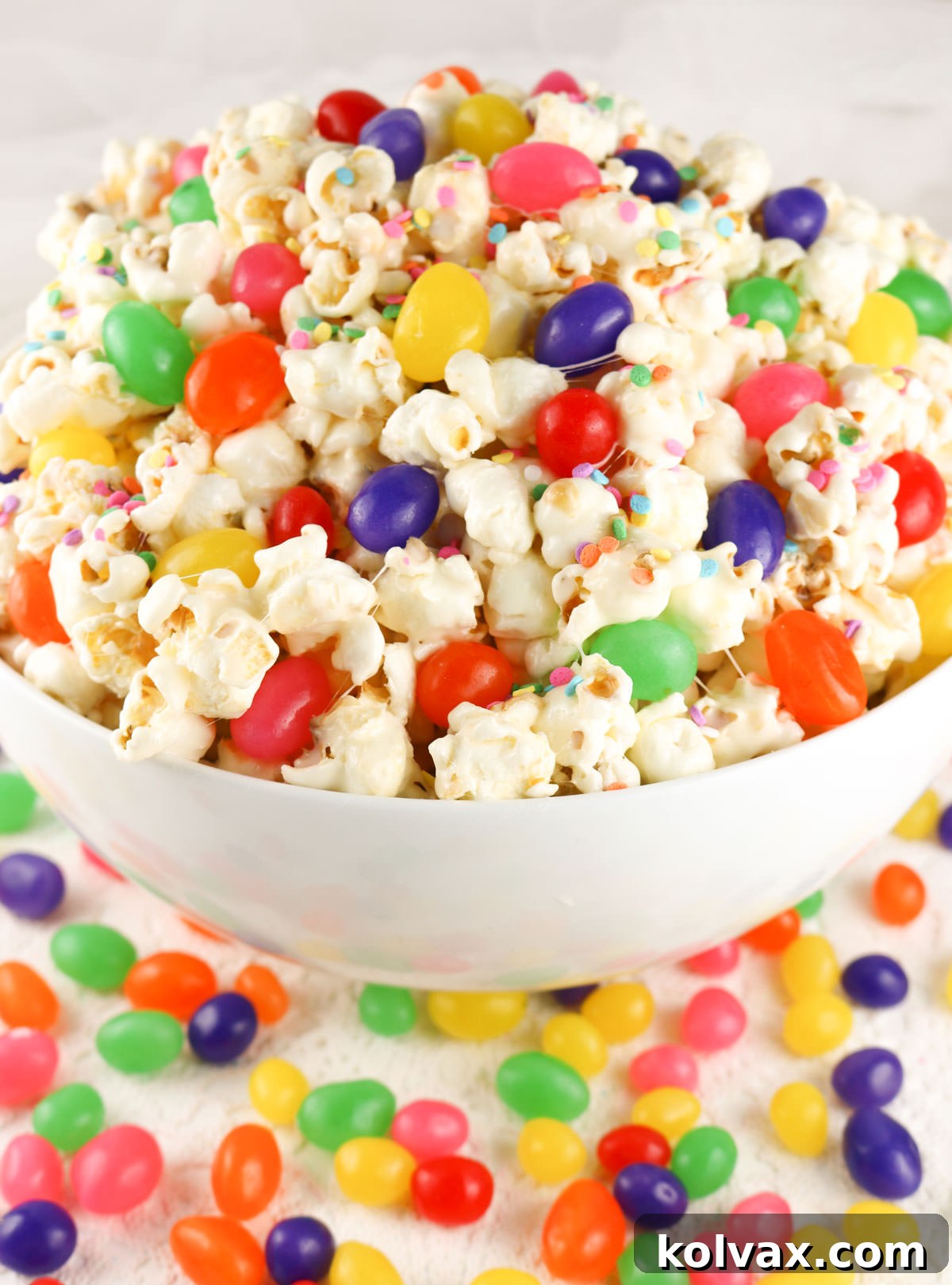 Closeup on a pristine white serving bowl generously filled with a vibrant, colorful mound of Jelly Bean Popcorn, surrounded by loose jelly beans on a white table, inviting guests to indulge in this sweet and salty Easter treat.